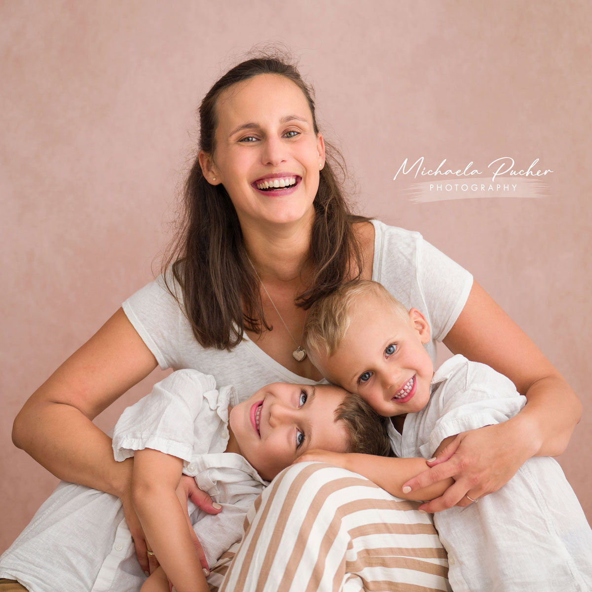 Mutter sitzt vor einem warmen, rosigen Hintergrund und hält ihre zwei fröhlich lächelnden kleinen Söhne im Arm, beide kuscheln sich eng an sie.